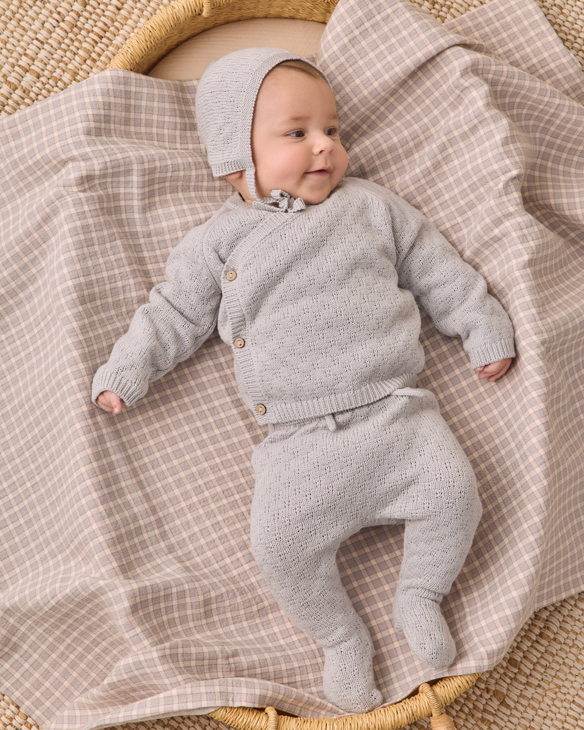 A baby wearing a light gray knit cardigan, matching footed pants, and a bonnet, lying on a beige and white checkered blanket in a woven basket.