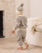 A baby wearing a matching ribbed knit two-piece outfit with a pom-pom beanie, shown from behind in a softly lit living room.