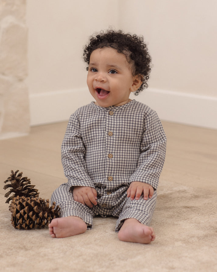 A baby with curly dark hair sits on a beige carpet next to a pinecone, wearing a long-sleeve button-front romper in a brown and beige gingham check pattern.