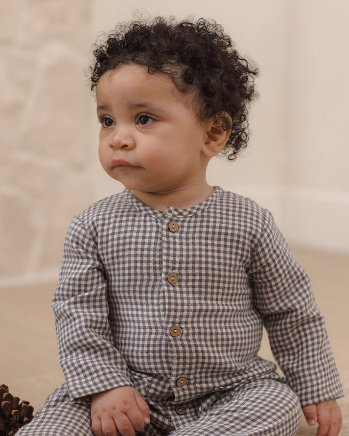 A baby with curly dark hair wearing a brown and beige gingham long-sleeve romper with brown buttons, sitting in a soft neutral indoor setting.