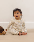 A baby sitting on a light wooden floor against a cream wall, wearing a cream-colored long-sleeve romper with a small blue floral pattern and a matching headband.