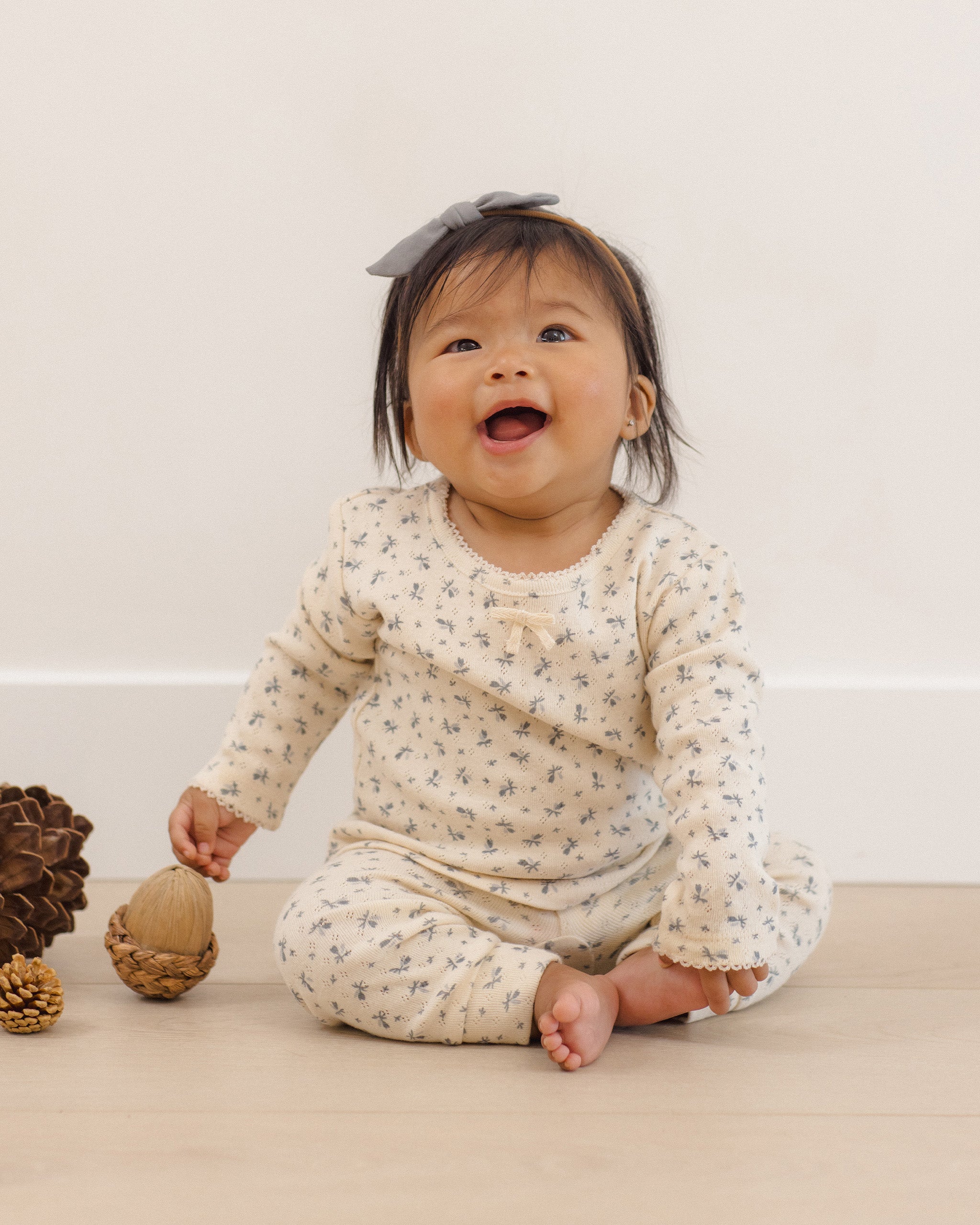 A baby sitting on a light wooden floor against a white wall, wearing a cream-colored long-sleeve romper with a small blue floral pattern and a gray bow headband.
