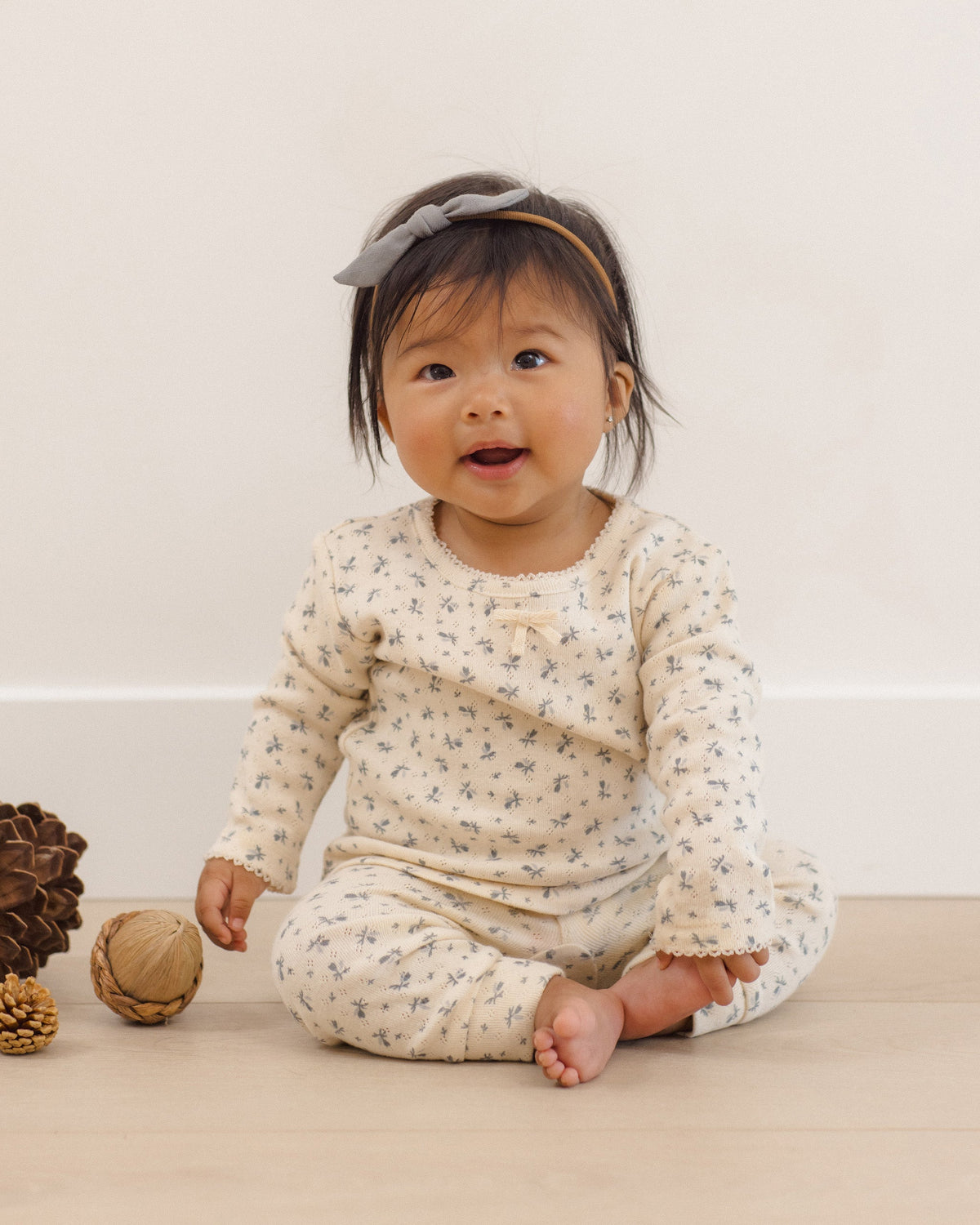 A baby sitting on a light wood floor wearing a cream-colored long-sleeve romper with a small blue floral pattern, a matching headband with a bow, and looking at the camera.