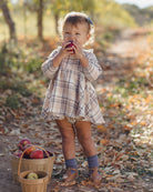 A toddler girl stands outdoors in a wooded path wearing a beige and brown plaid dress, eating an apple. A small wooden basket filled with apples sits beside her.