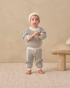 A baby standing barefoot on a textured beige rug, wearing a cream knit beanie and a matching cream and gray striped knit two-piece outfit. The background is a solid beige wall with a rustic wooden mushroom-shaped stool on the right.