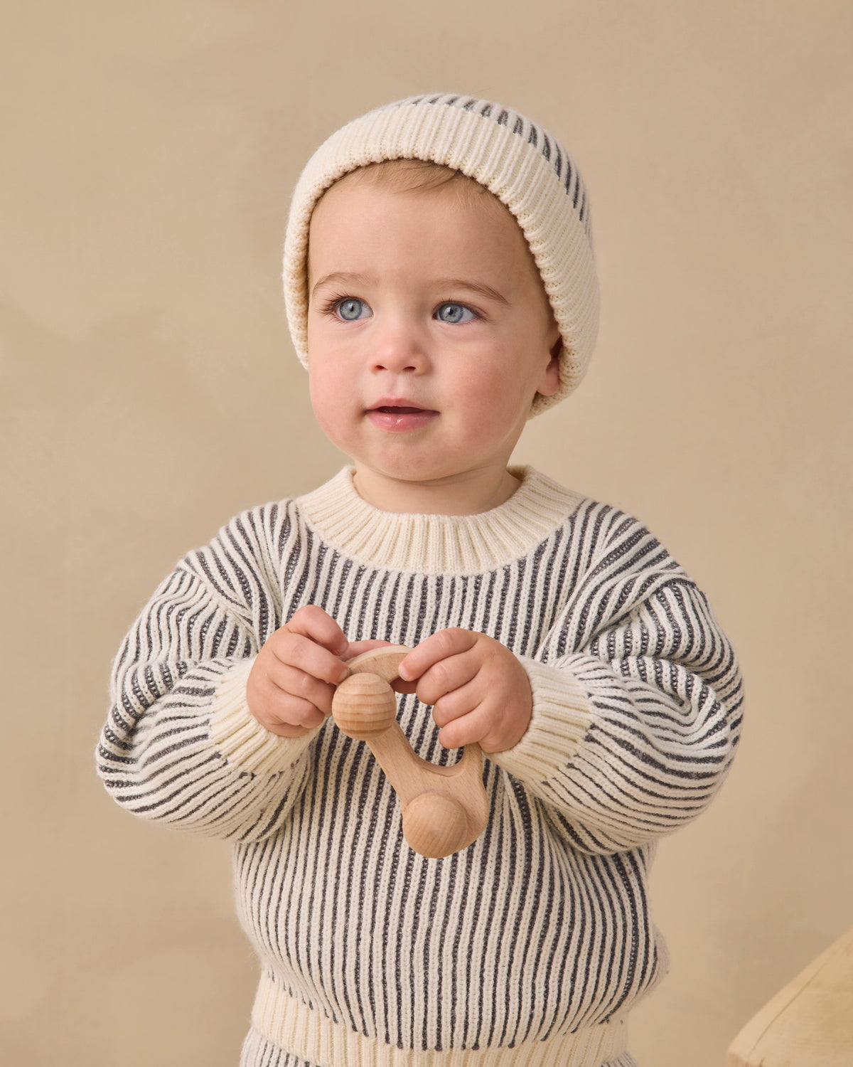 A baby wearing a cream and navy striped knit sweater and matching beanie hat, holding a wooden teething toy against a warm beige background.