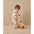 A toddler stands barefoot on a textured cream rug against a warm beige wall, wearing a cream long-sleeve romper with small blue embroidered star-like motifs. The child is holding dried grass near a small woven basket.