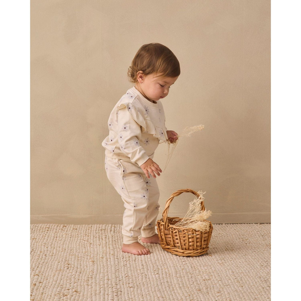 A toddler stands barefoot on a textured cream rug against a warm beige wall, wearing a cream long-sleeve romper with small blue embroidered star-like motifs. The child is holding dried grass near a small woven basket.