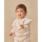 A toddler girl wearing a cream-colored two-piece outfit with small dark polka dots, standing against a solid beige background and holding a small sprig of dried flowers.