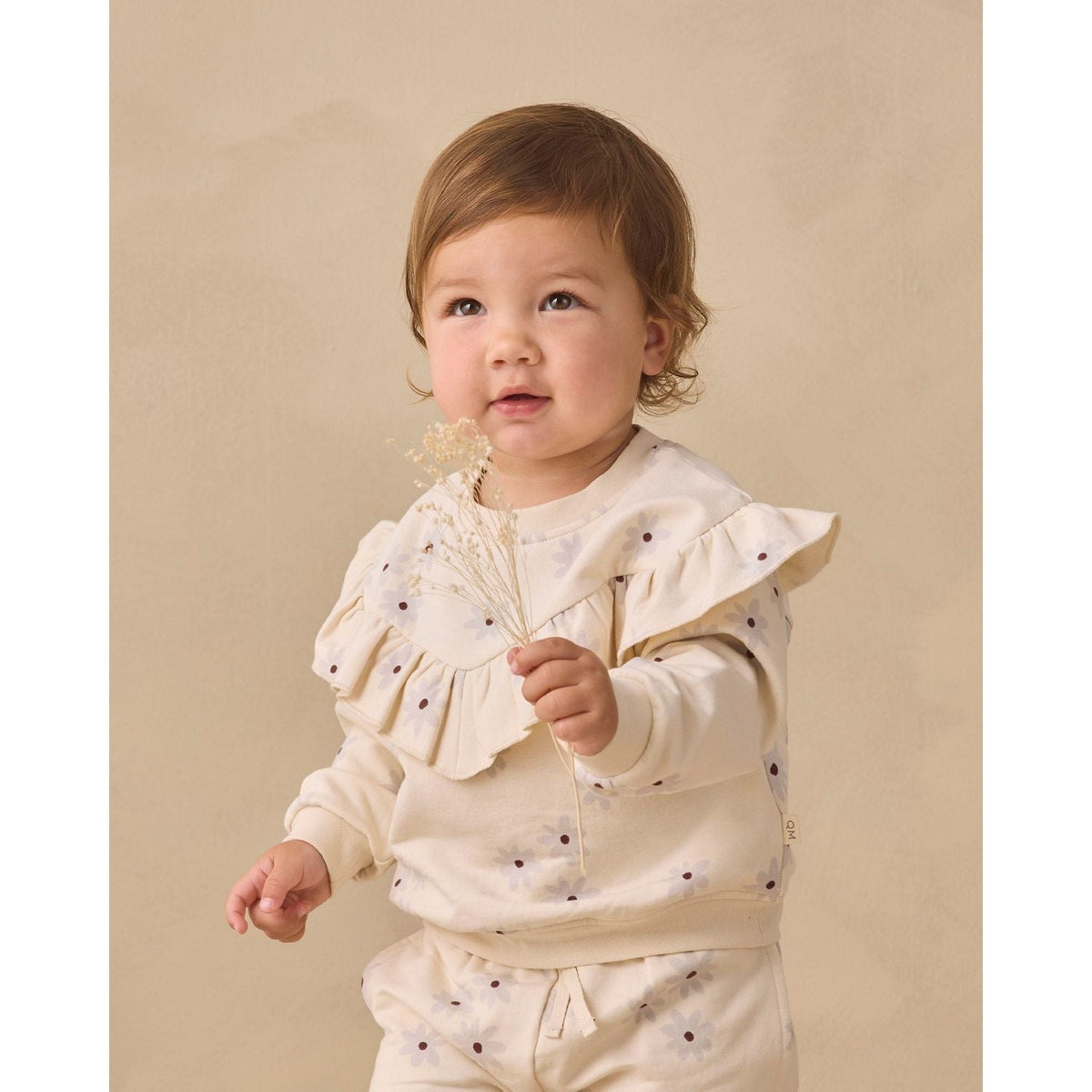 A toddler girl wearing a cream-colored two-piece outfit with small dark polka dots, standing against a solid beige background and holding a small sprig of dried flowers.