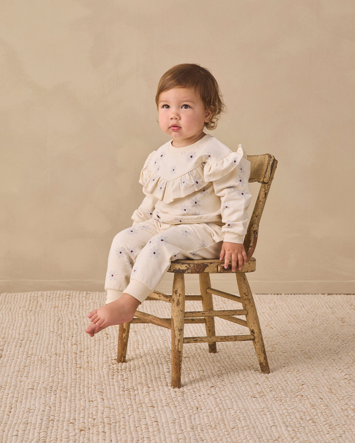 A toddler sits on a small rustic wooden chair wearing a cream ruffled long-sleeve top and matching pants with tiny dark blue star prints, set against a warm beige studio backdrop.