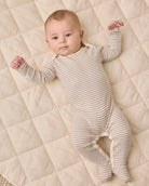 A baby lying on a quilted cream blanket, wearing a long-sleeve footed onesie with thin horizontal gray stripes.