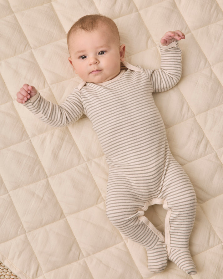 A baby lying on a quilted cream blanket, wearing a long-sleeve footed onesie with thin horizontal gray stripes.
