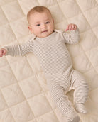 A baby lying on a quilted cream mattress, wearing a long-sleeve, footed one-piece romper with thin horizontal stripes in beige and white.