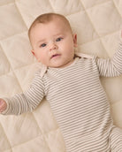A baby lying on a quilted cream mattress, wearing a beige and white horizontally striped long-sleeve bodysuit.