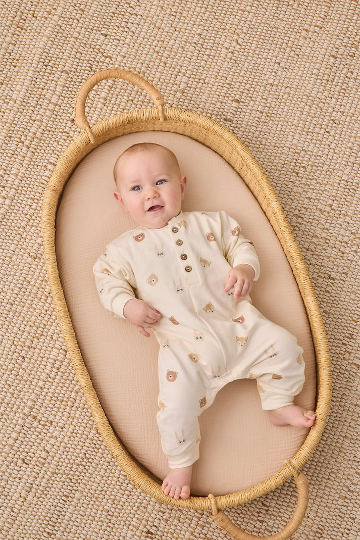 A baby lying on their back inside a woven oval bassinet, wearing a cream-colored footless onesie with small brown animal prints. The bassinet is placed on a textured beige woven rug.