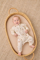 A baby lying on their back inside a woven oval bassinet, wearing a cream-colored footless onesie with small brown animal prints. The bassinet is placed on a textured beige woven rug.