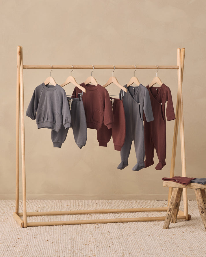 A wooden clothing rack displaying five children's matching sleepwear sets in gray and burgundy, styled in a minimalist studio setting with a small wooden stool.