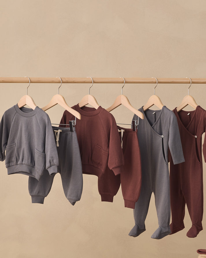 Five children's two-piece clothing sets hanging on wooden hangers from a wooden rod against a warm beige background. The sets include gray sweatshirts and pants, a dark brown sweatshirt and pants, and a gray wrap-style sweatshirt and footed pants, with one dark brown footed sweatshirt set on the far right.