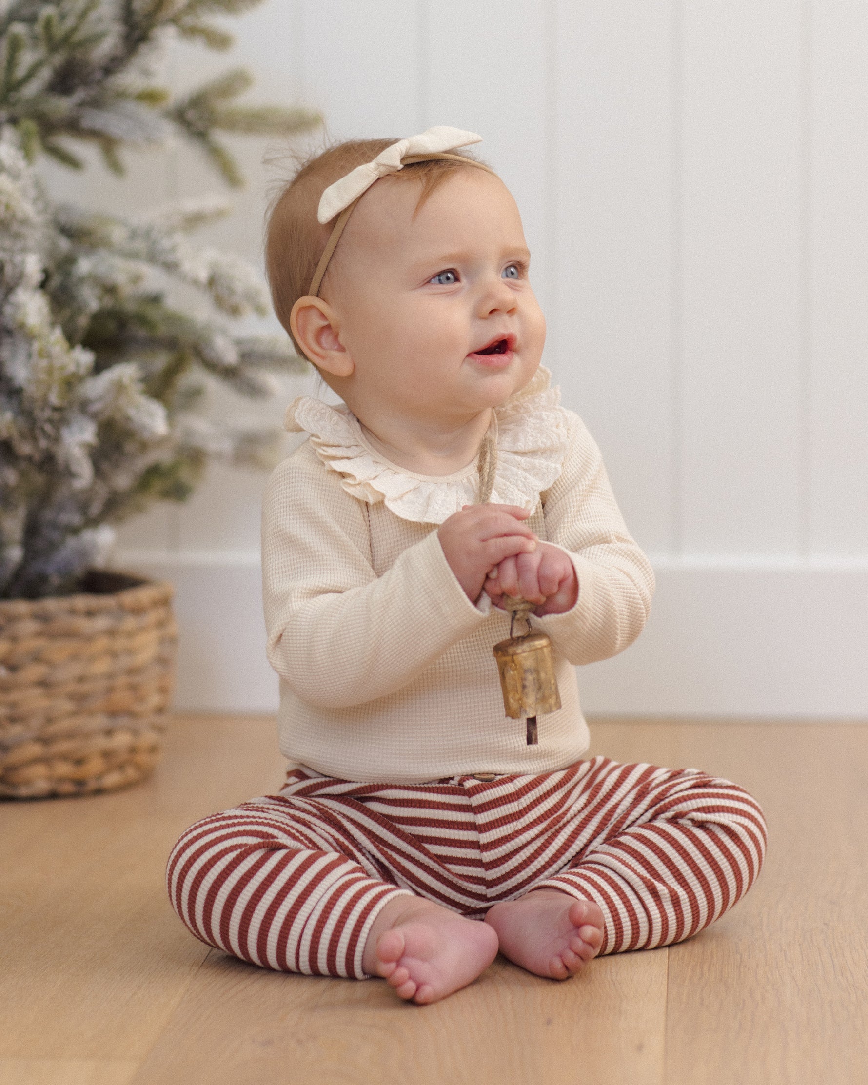 A baby sitting on a wooden floor, wearing a long-sleeve natural beige ruffled collar bodysuit and burgundy-and-white striped pants, holding a small brass bell while wearing a headband with a bow.