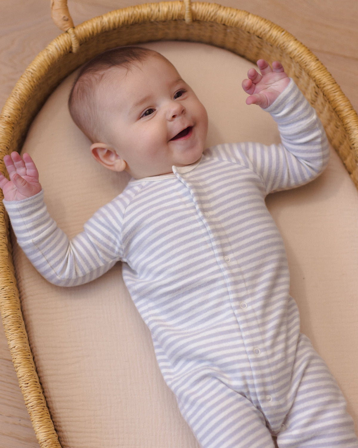 A smiling baby lying inside a round wicker basket, wearing a soft, organic cotton long-sleeve romper in a white and gray horizontal stripe pattern by Quincy Mae.