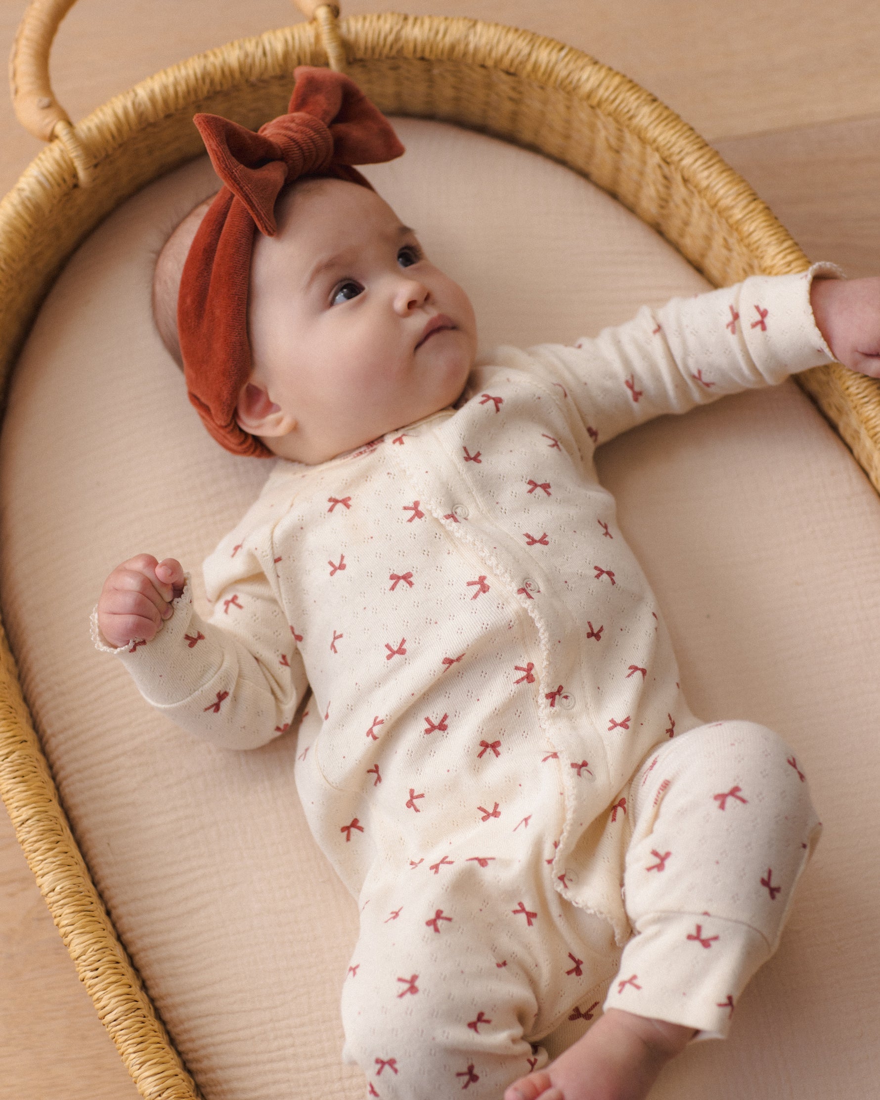 Baby wearing a redwood velour bow headband and a patterned cream-colored outfit, lying in a wicker basket.