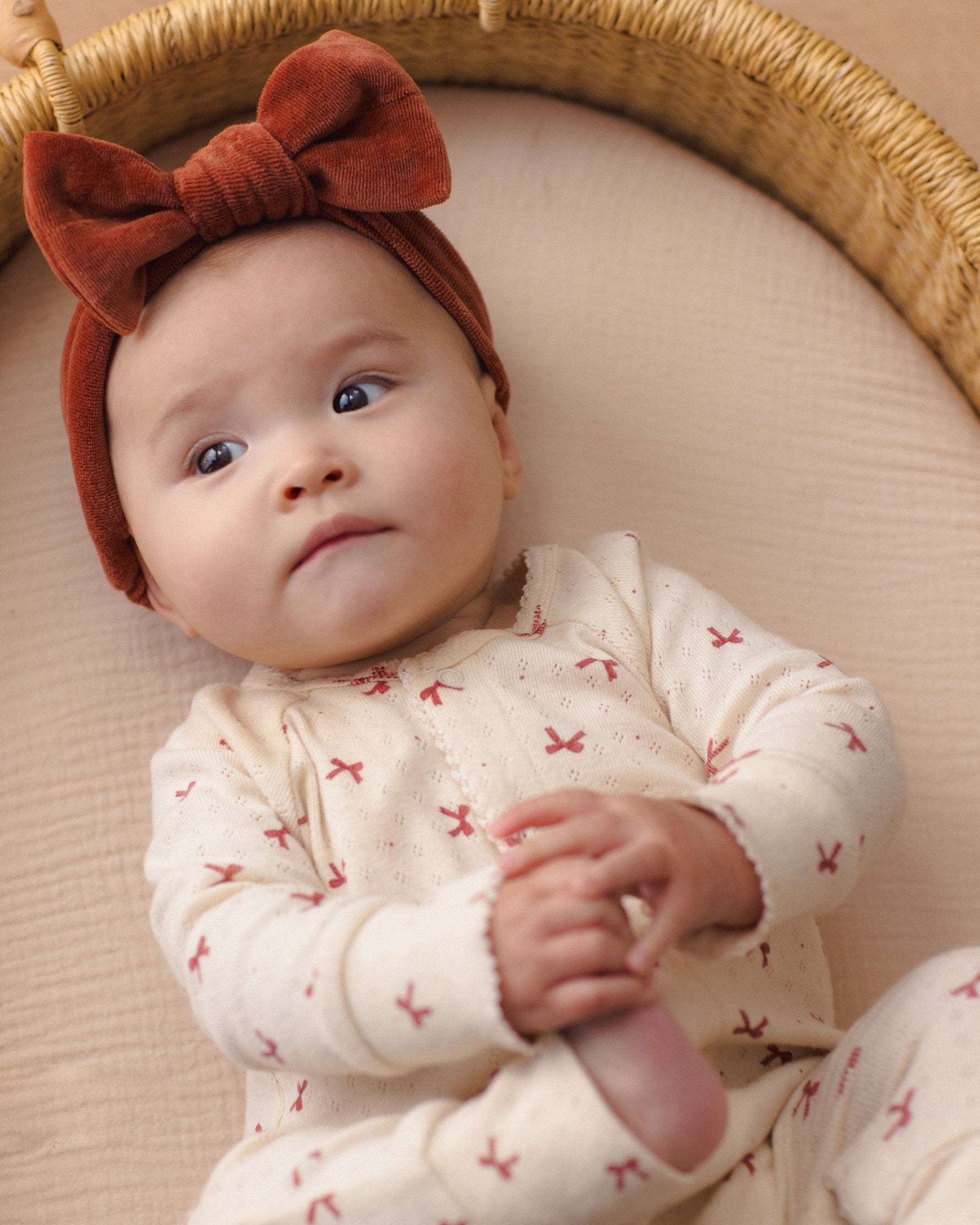 Baby wearing a redwood velour bow headband and a patterned cream-colored outfit, lying in a wicker basket.