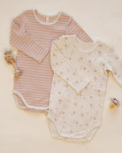 Two long-sleeve baby bodysuits laid flat on a warm cream background. The bodysuit on the left is light pink and white striped, and the bodysuit on the right is cream with a small pink floral print.