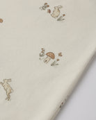 A close-up of cream-colored fabric printed with a whimsical pattern of small rabbits, mushrooms, and flowers in muted brown and olive tones.