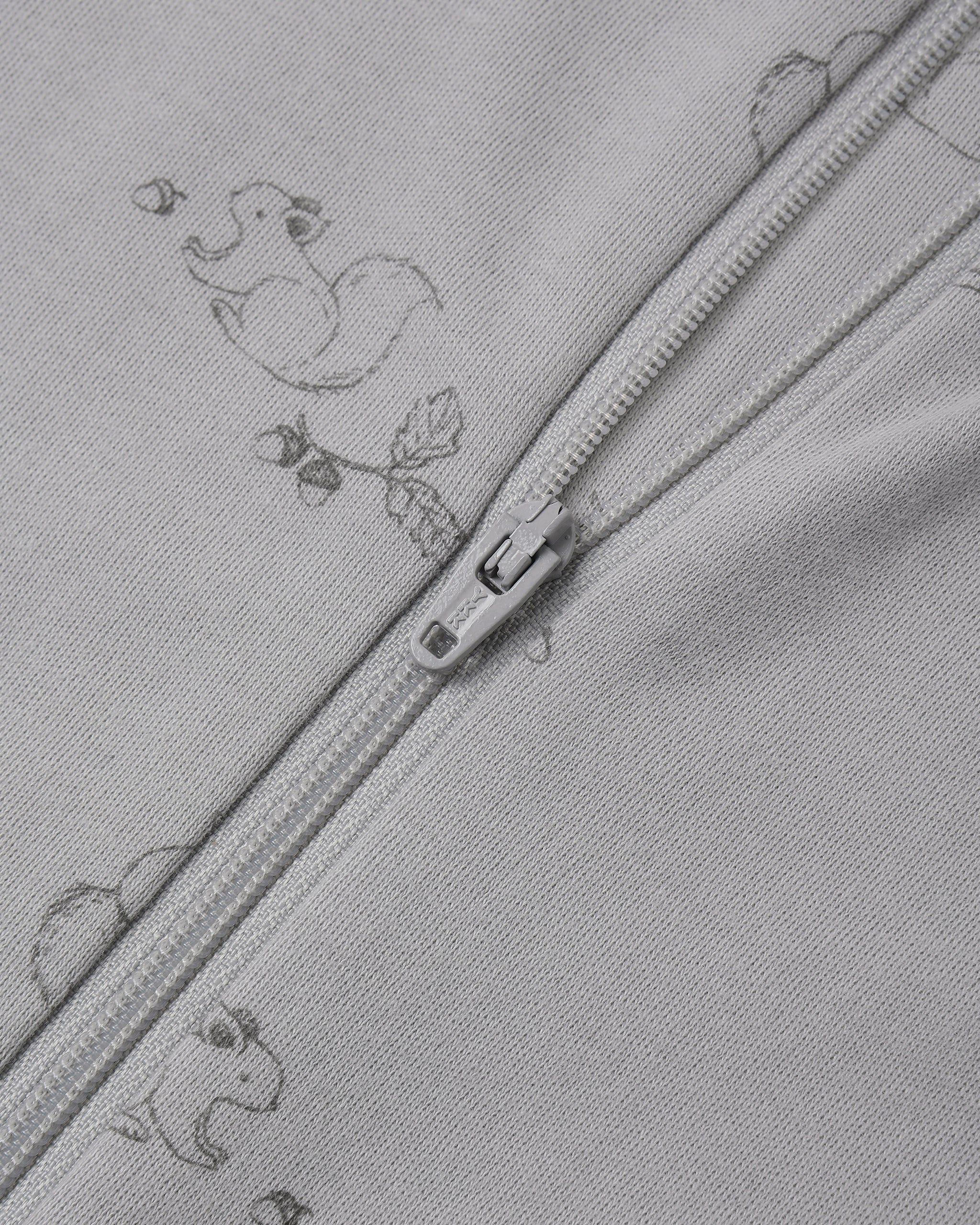 Close-up of a light gray zippered garment with a subtle repeating duck and leaf print, showing the zipper pull and fabric texture.
