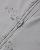 Close-up of a light gray zippered garment with a subtle repeating duck and leaf print, showing the zipper pull and fabric texture.