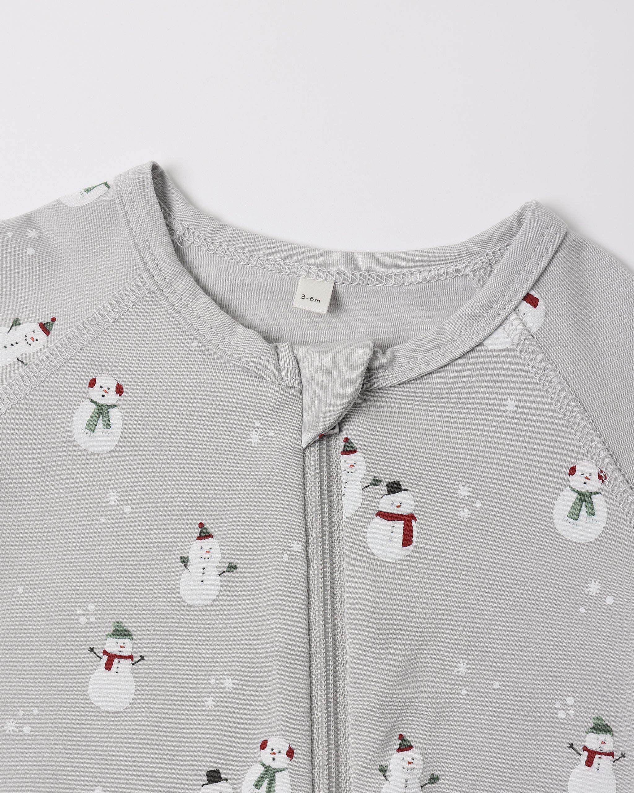 A close-up view of the upper chest and collar area of a light grey Quincy Mae bamboo zip footie, featuring an all-over wintery snowman print and a central zipper.
