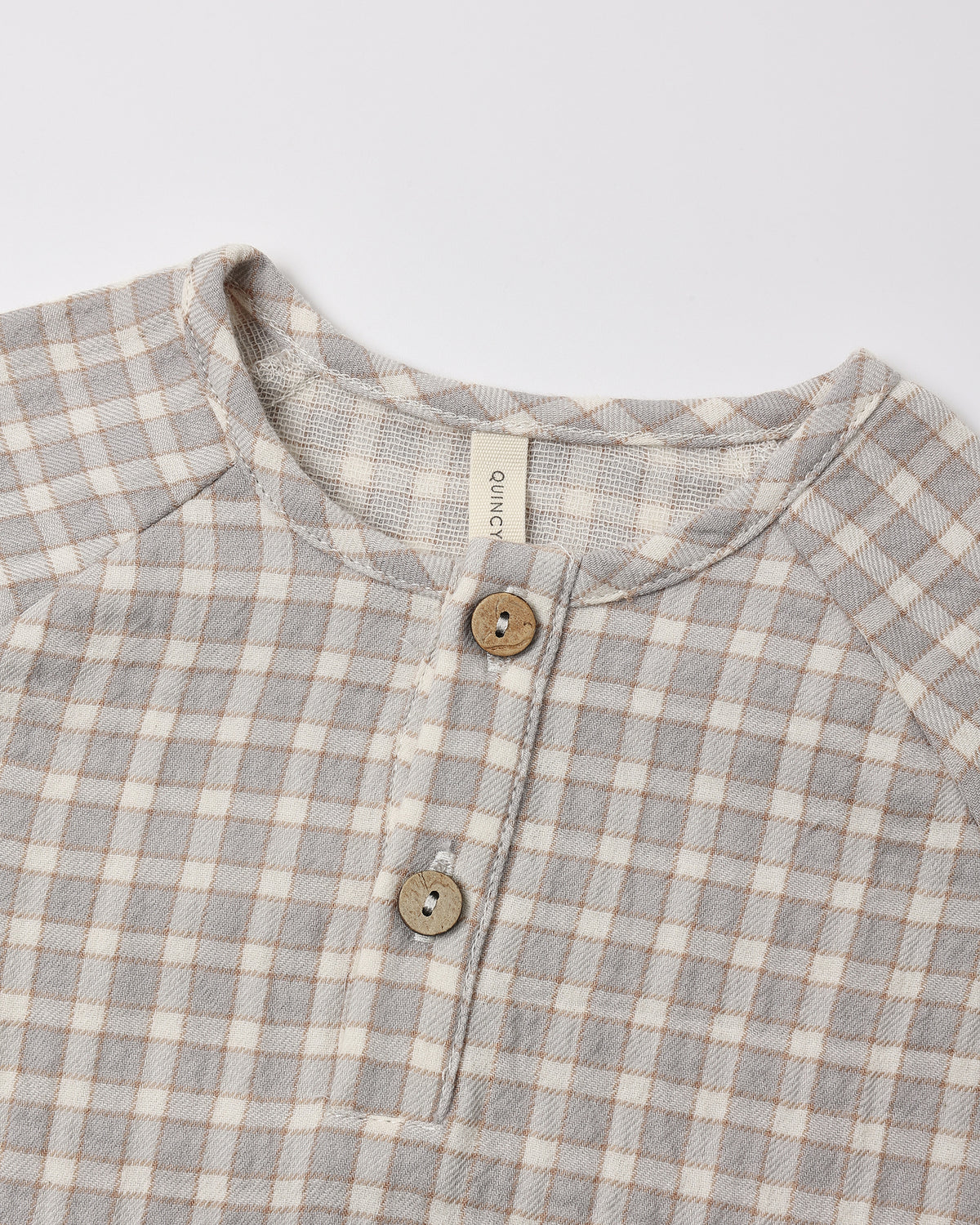 Close-up of a children's long-sleeve henley shirt with a light gray and beige gingham check pattern, featuring a button placket with wooden buttons and a visible neck label reading 'gugu'.