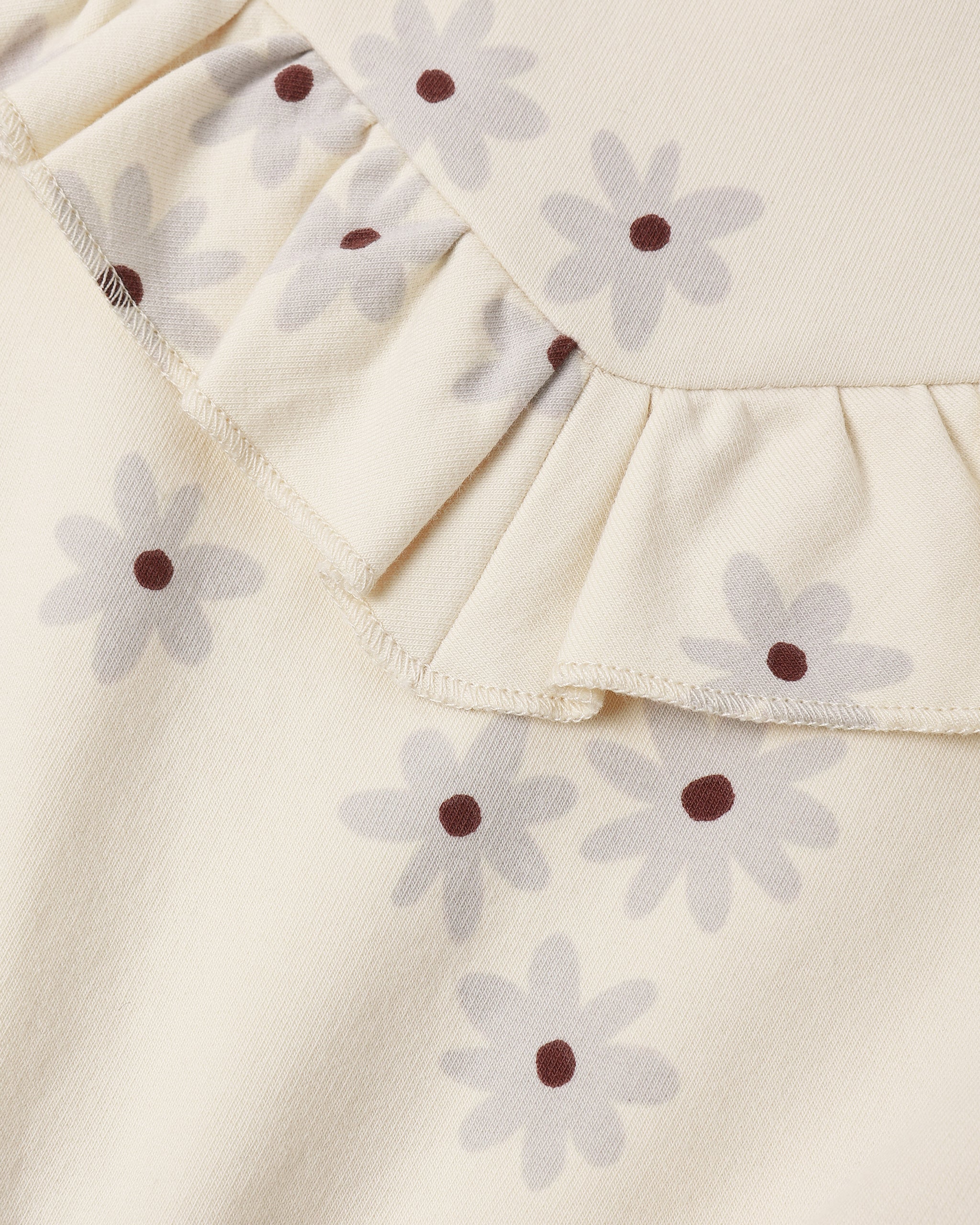 Close-up of a cream-colored fabric garment featuring a ruffled neckline and an all-over daisy print with light gray petals and dark brown centers.