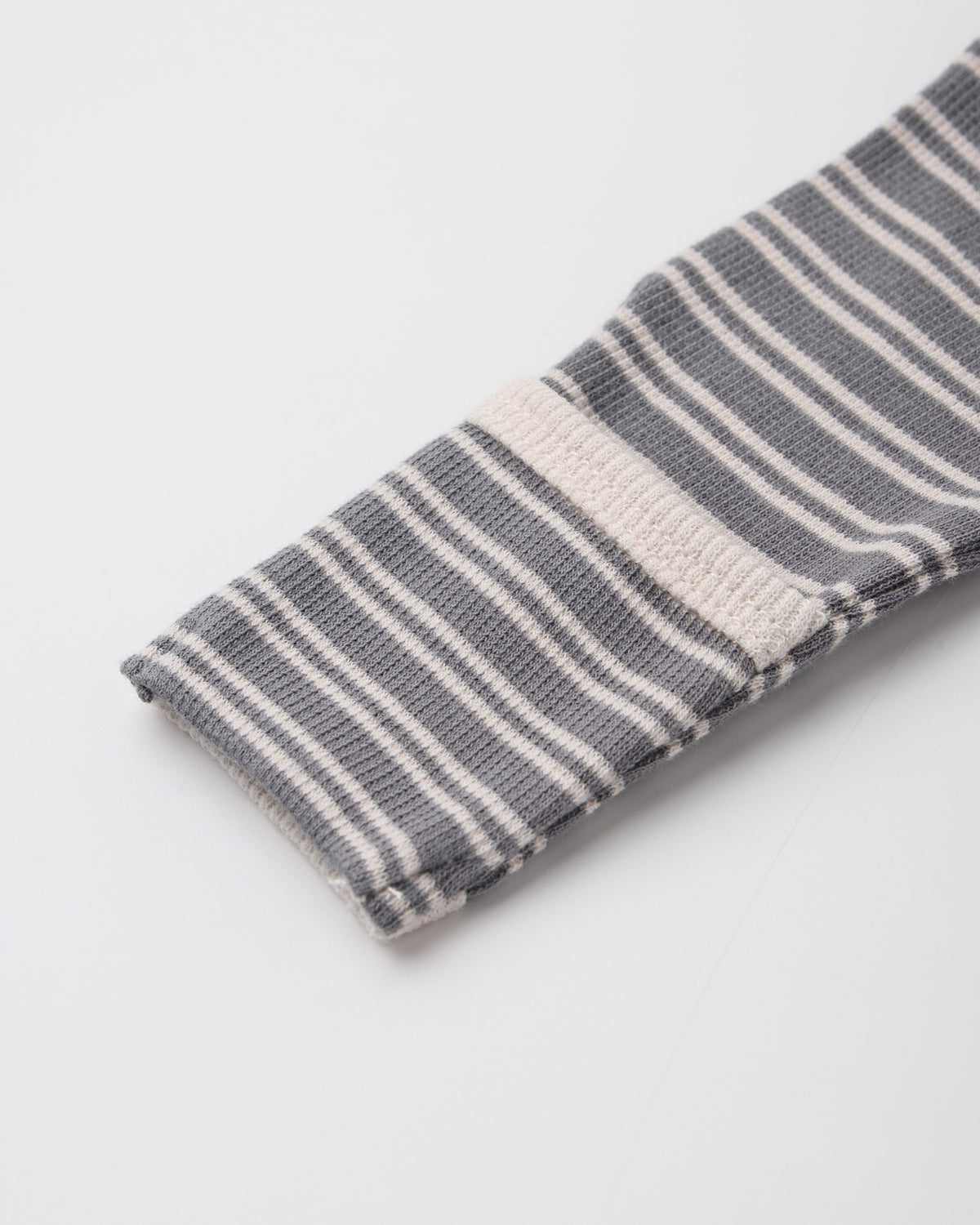 A close-up of a folded knit fabric item, likely a sock or sleeve cuff, featuring a gray and white striped pattern with a solid white stripe accent, on a light gray background.