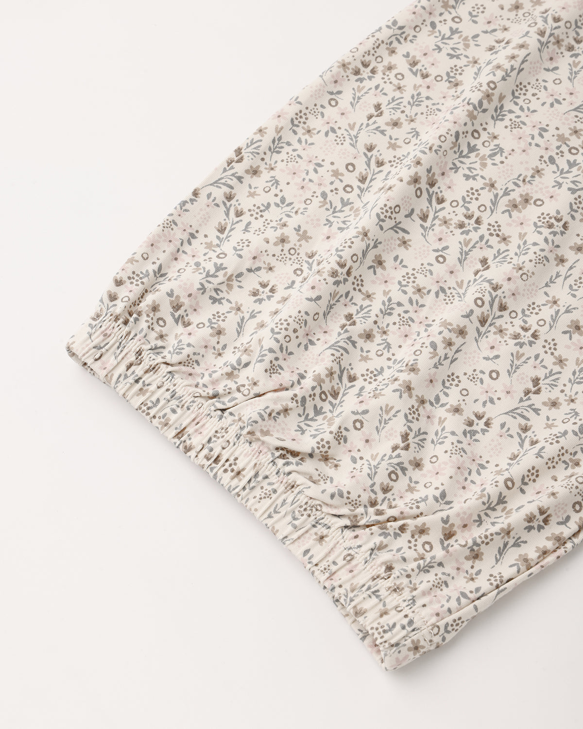 A close-up of a lightweight fabric with a delicate, small-scale floral and botanical print in muted gray and brown tones on a warm cream background, finished with an elasticized ruffled hem.