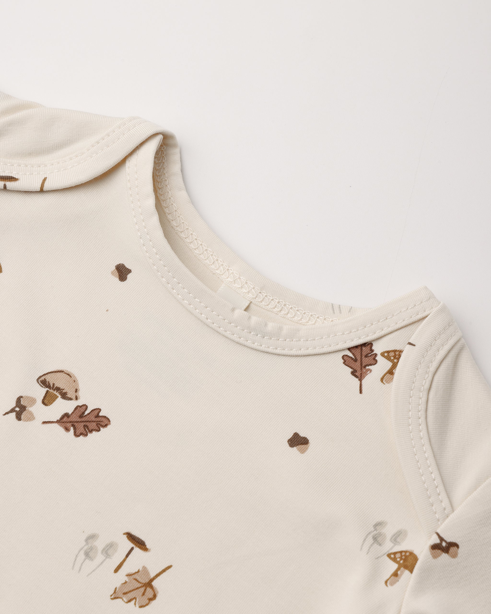 Close-up of a cream-colored baby top with a delicate brown and beige autumn leaf and mushroom print, showing the ribbed collar and shoulder seams.