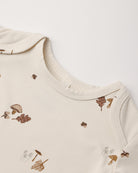 Close-up of a cream-colored baby top with a delicate brown and beige autumn leaf and mushroom print, showing the ribbed collar and shoulder seams.