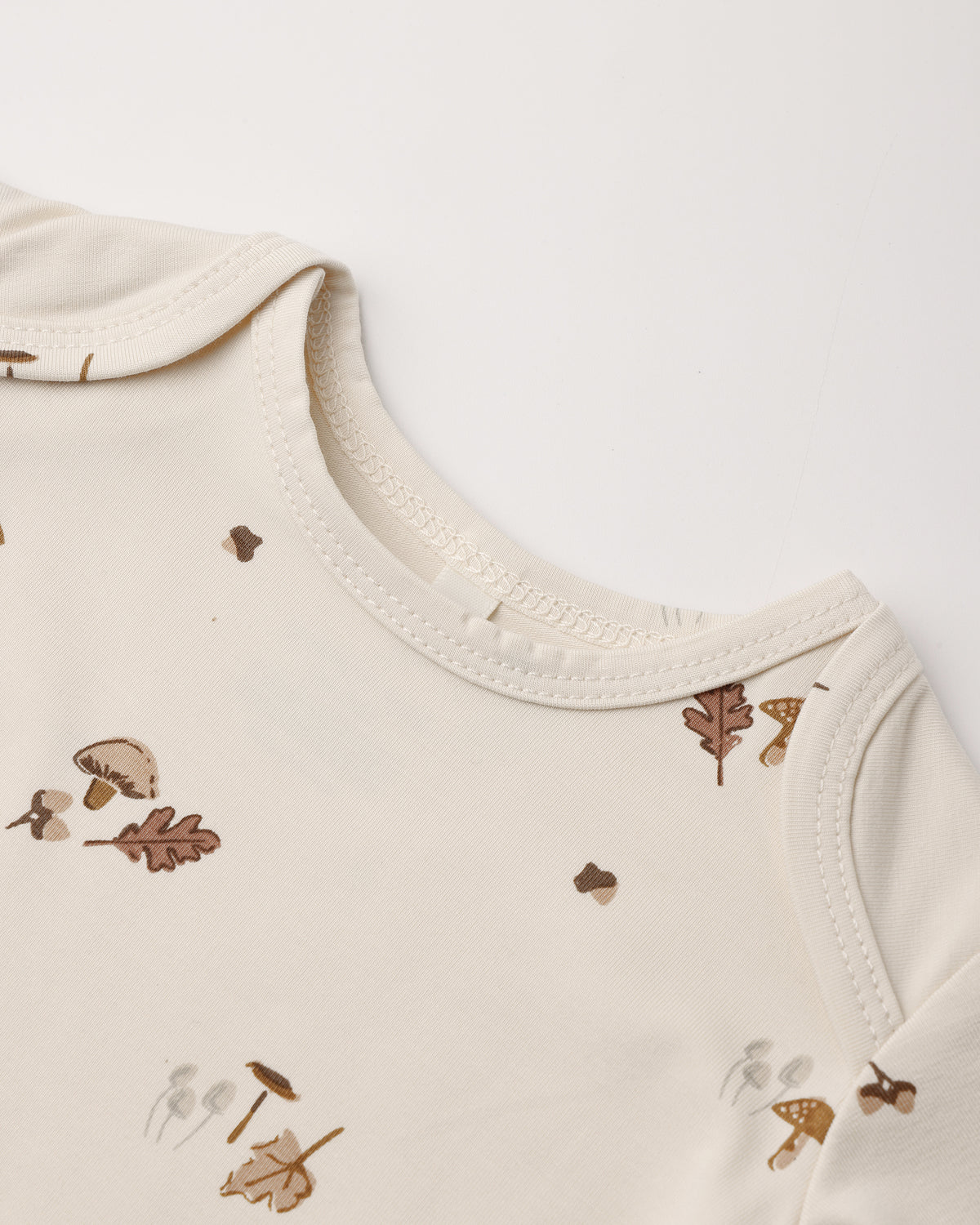 Close-up of a cream-colored baby top with a delicate brown and beige autumn leaf and mushroom print, showing the ribbed collar and shoulder seams.