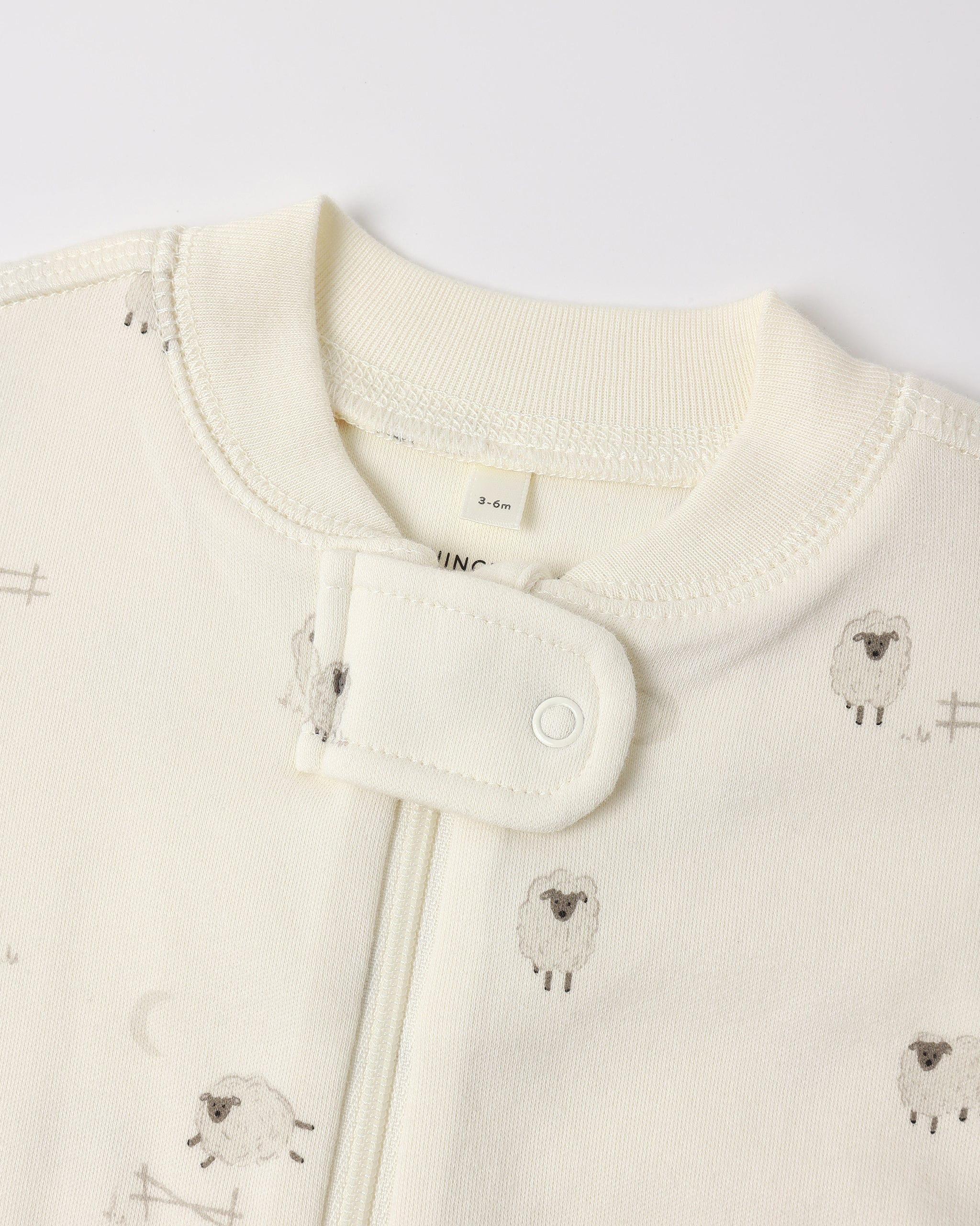 Close-up of a baby garment in warm off-white/cream with a small sheep print and a snap-button closure at the neckline.