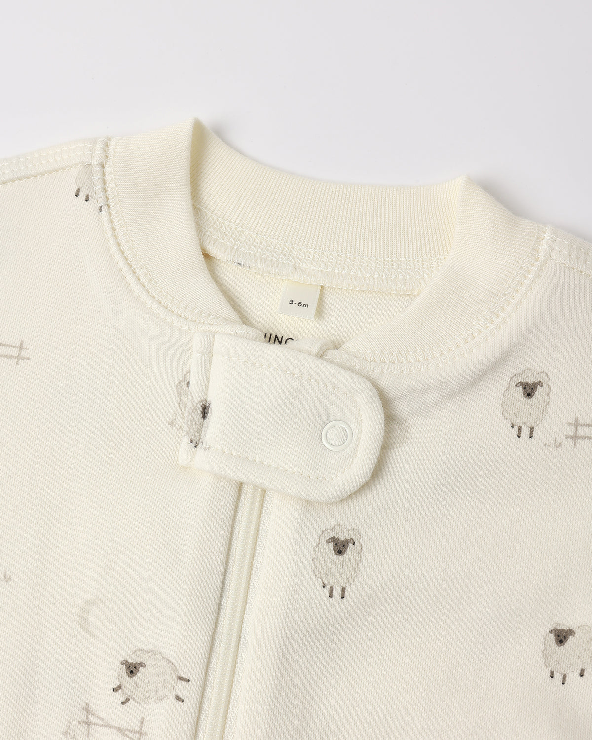 Close-up of a baby garment in warm off-white/cream with a small sheep print and a snap-button closure at the neckline.