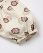 A close-up of a cream-colored fabric garment, likely a baby romper or skirt, featuring an all-over floral print in dusty mauve and beige, finished with a gathered elastic cuff.