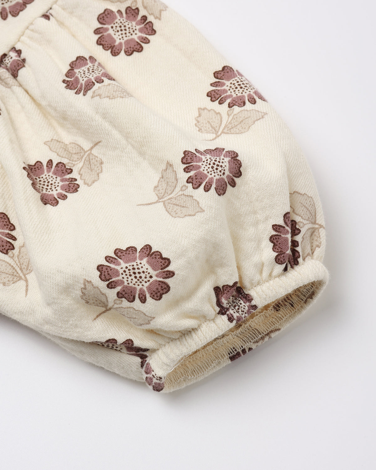 A close-up of a cream-colored fabric garment, likely a baby romper or skirt, featuring an all-over floral print in dusty mauve and beige, finished with a gathered elastic cuff.