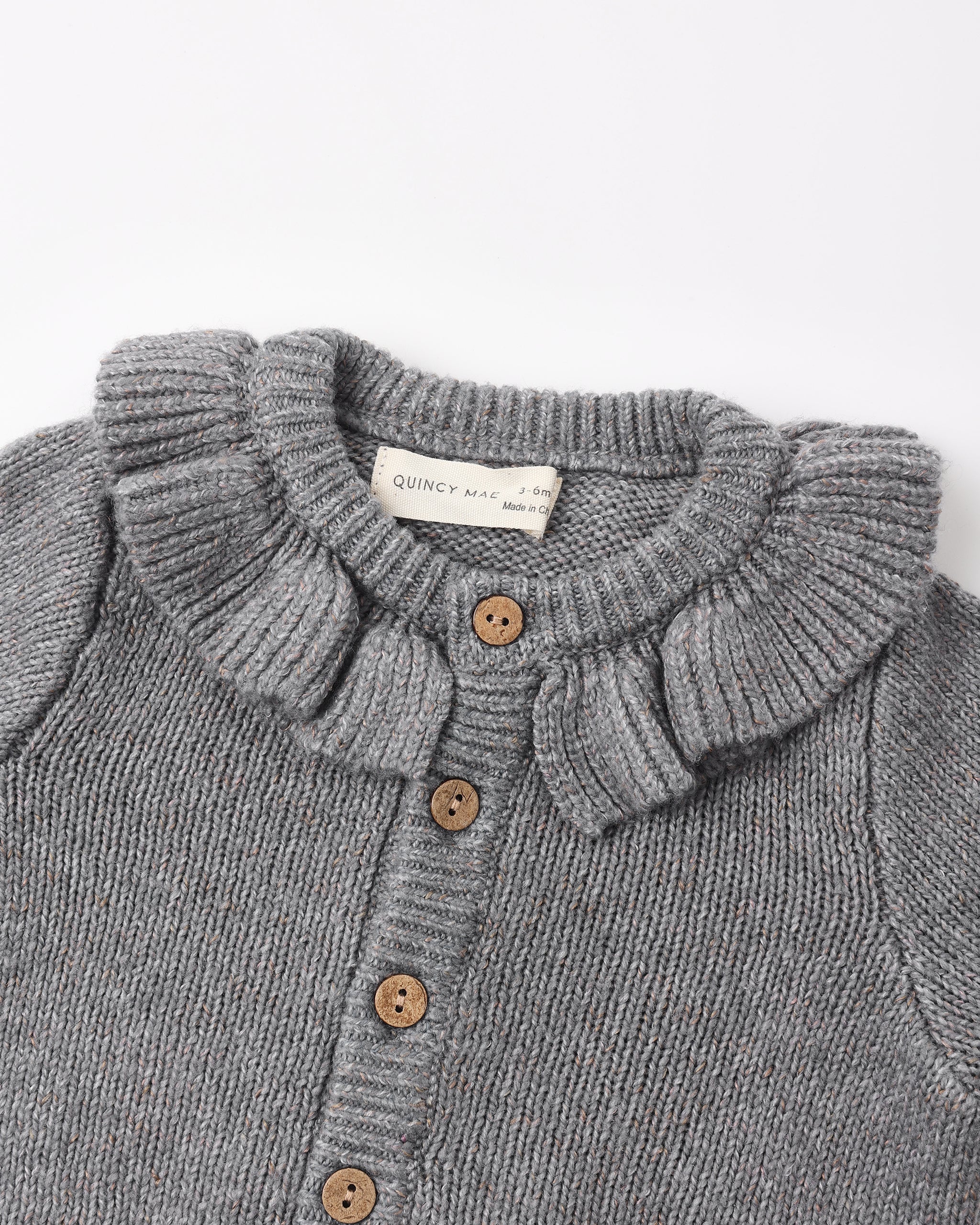 Close-up of a gray ribbed knit cardigan sweater with a ruffled Peter Pan collar and brown buttons, showing a brand label that reads 'QUINCY MART'.