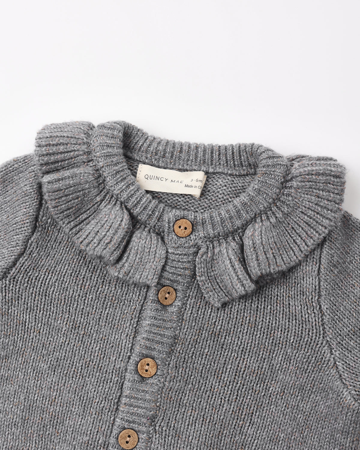 Close-up of a gray ribbed knit cardigan sweater with a ruffled Peter Pan collar and brown buttons, showing a brand label that reads 'QUINCY MART'.