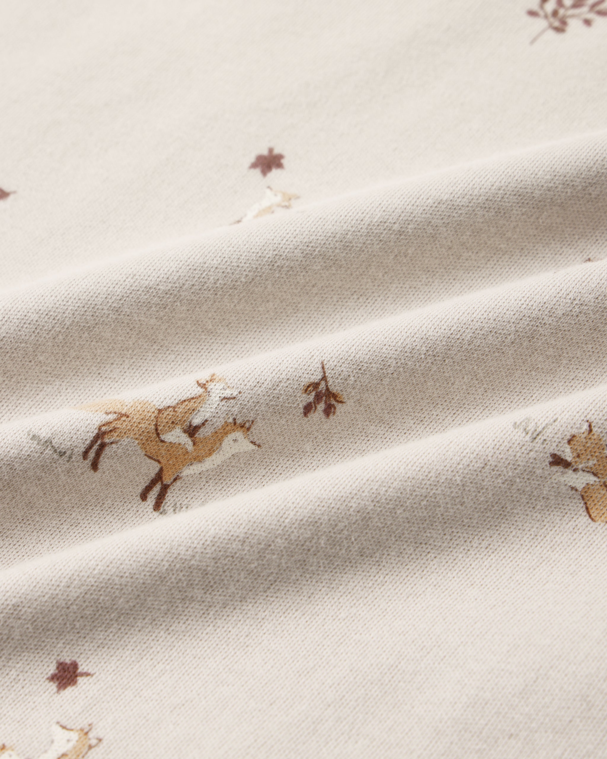 A close-up of cream-colored fabric with a printed pattern of brown deer and small brown leaves, shown with soft folds.