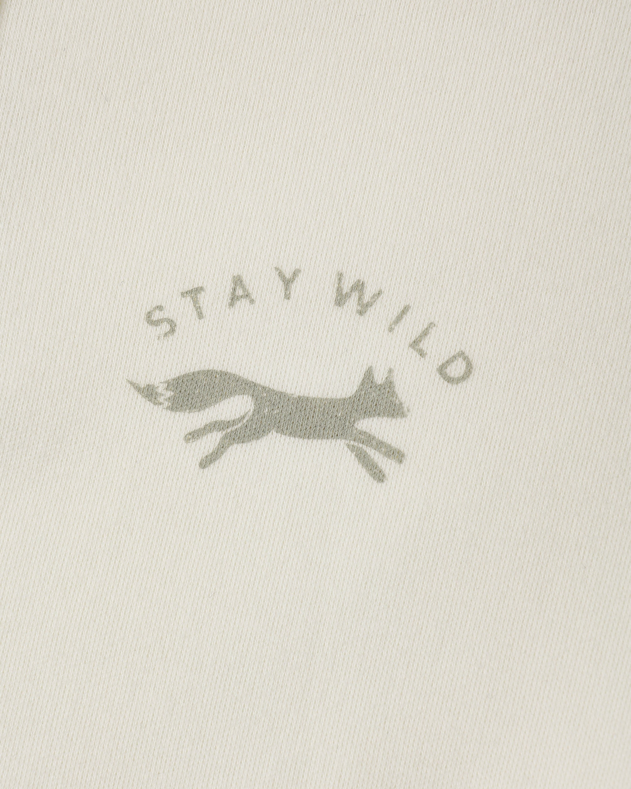 Close-up of a light cream fabric with a muted gray printed logo featuring the words 'STAY WILD' and a running fox silhouette.
