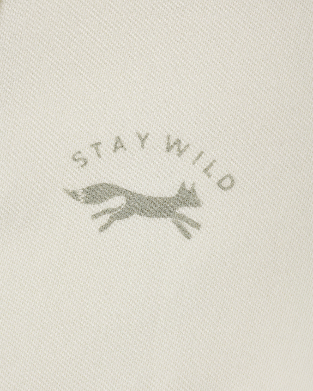 Close-up of a light cream fabric with a muted gray printed logo featuring the words 'STAY WILD' and a running fox silhouette.