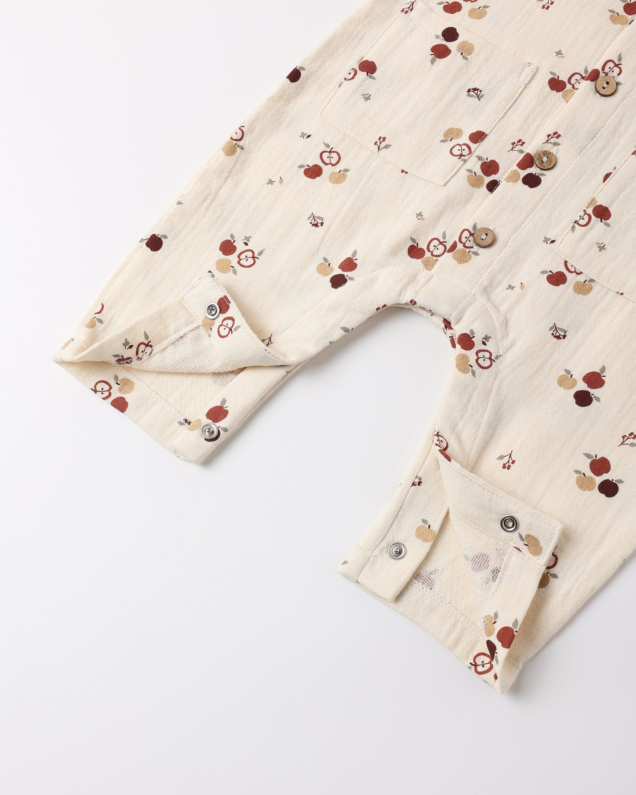 A close-up of a cream-colored cotton gauze baby romper or jumpsuit with a small red, brown, and beige fruit print, featuring a buttoned cuffed sleeve and a chest pocket.