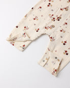 A close-up of a cream-colored cotton gauze baby romper or jumpsuit with a small red, brown, and beige fruit print, featuring a buttoned cuffed sleeve and a chest pocket.